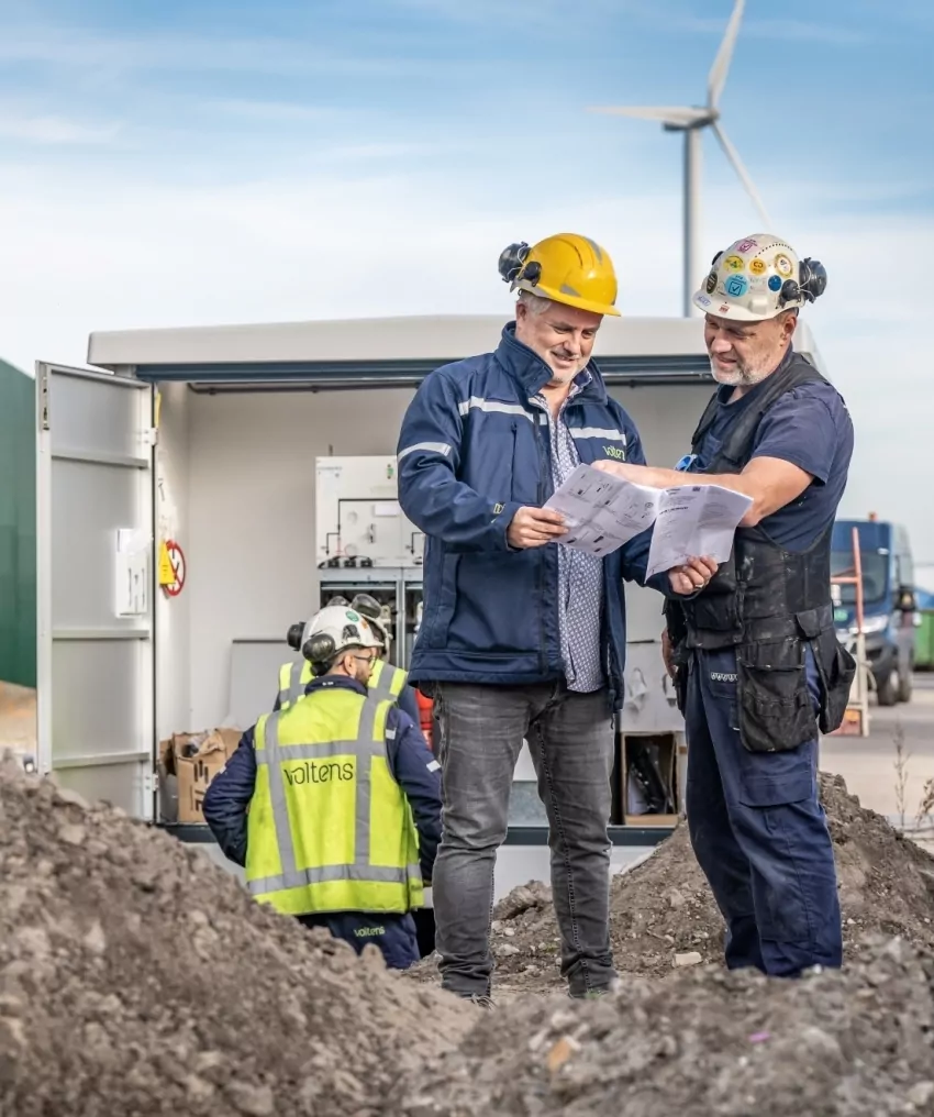 Twee mannen met bouwhelm bekijken een tekening bij een geopend transformatorstation