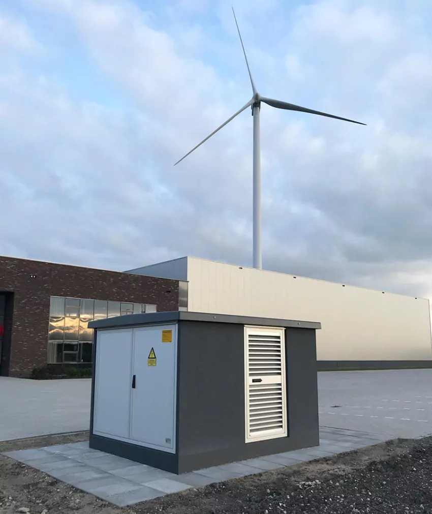 Wit transformatorstation in een duinlandschap met een windturbine erachter